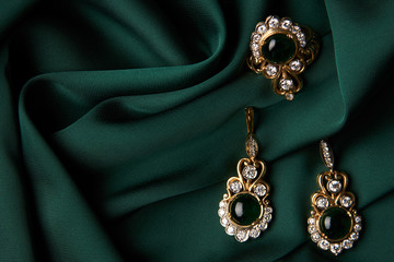 Beautiful Golden ring and pair of earrings with green Emerald and Diamonds gemstones on a green satin background. Luxury female jewelry, close-up. Selective focus