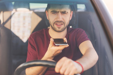 Serious concentrated unshaven brunet male drives car, makes voice call, speaks with colleague, goes for work, stands in traffic jam, being busy. Handsome man with modern cell phone in automobile © sementsova321