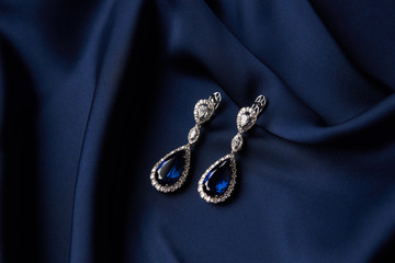 Two Golden sapphire earrings with small diamonds. Pair of platinum earring with sapphire gemstone on blue satin background. Luxury female jewelry, close-up