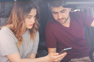 Photo of concentrated girlfriend and boyfriend focused in mobile phone, watch video online, sit together in automobile, being in traffic jam. People, travelling. Two female and male passengers