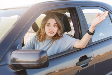Photo of puzzled woman gestures though car window, being annoyed to wait for long and stand in traffic jam, hurries for important meeting or work. People, driving, transport and emotions concept
