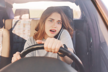 Surprised young cute female driver has telephone conversation via cell phone, drives car, looks with shocked expression as forgets about something important or hears unexpected news from friend.