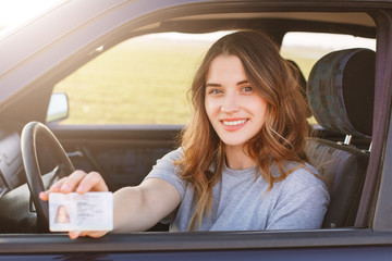 Smiling young female with pleasant appearance shows proudly her drivers license, sits in new car,...