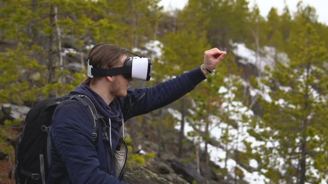 The artist-tourist in glasses of virtual reality draws a picture. In the background there is a coniferous forest and snowy mountain slopes.