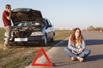 Helpless couple stop on road as have breakdown of car. Desperate male calls somebody via cell phone, asks for help while stands near brocken transport, thoughtful female sits on asphalt being in panic
