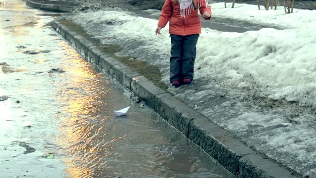 Girl in rubber boots jumping on puddle near wite paper boat