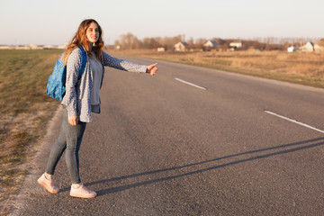 Outdoor shot of pretty smiling female traveler stands on road, hitchhikes and stops cars, carries bag, enjoy free lifestyle, has positive expression. Woman hitchhiker stands on country road alone