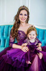 Beautiful mother and daughter in luxurious purple dress and tiara, sitting on the couch