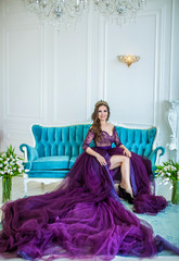Fototapeta premium Beautiful princess in purple fluffy long dress and with diadem sits on luxurious blue couch