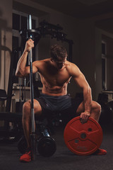 Fototapeta na wymiar Muscular shirtless athlete holds barbell disc while sits on a bench in the gym.