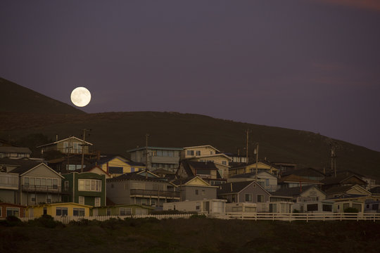 Full moon rising above a small beach town at the base of a hill at dusk.