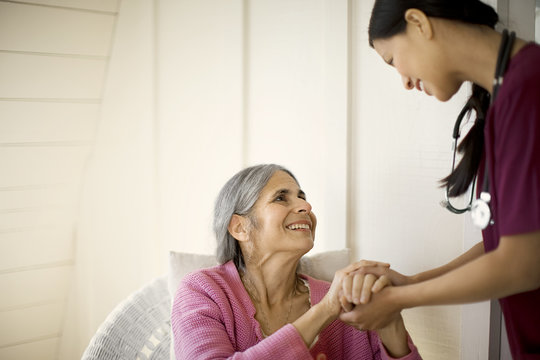 Smiling senior woman holding hands and speaking with a female nurse while seated in a chair.