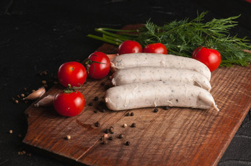 Three raw sausages on a wooden brown board on a black background with cherry tomatoes, parsley and dill, pepper and garlic.