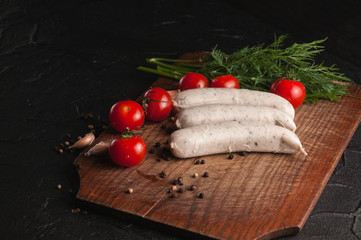 Three raw sausages on a wooden brown board on a black background with cherry tomatoes, parsley and dill, pepper and garlic.