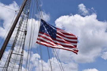 American flag with 15 stars waving from tall ship mast, blue sky with clouds.