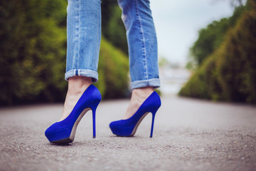girl in jeans stands on high blue heels on the road in the park in the summer