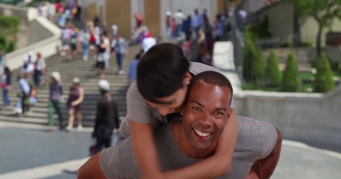 Young African man spinning around with his pretty girlfriend on his back near the Spanish Steps in Rome, Cute young couple playfully spending time together outside in Rome, 4k