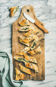Flat-lay of fresh Turkish borek roll cut in slices slices with spinach, feta cheese, black cumin seeds on wooden board over grey marble background, top view. Traditional East Mediterranean cuisine