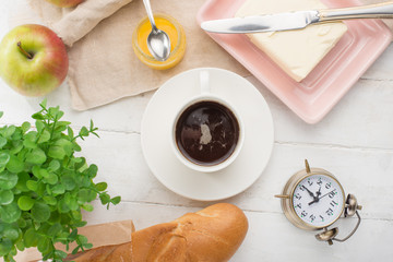 Morning cup of coffee, alarm clock, apples, butter and baguette, in a light kitchen. Background area, the concept of a bright morning and breakfast. View from above