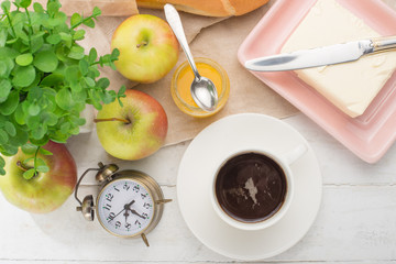 Morning cup of coffee, alarm clock, apples, butter and baguette, in a light kitchen. Background area, the concept of a bright morning and breakfast. View from above