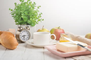 Morning cup of coffee, alarm clock, apples, butter and baguette, in a light kitchen. Background...