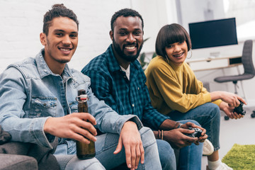 mixed race man with bottle of beer and smiling friends with joysticks behind