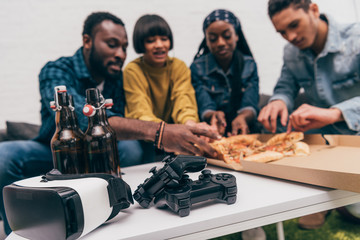 closeup shot joysticks, bottles of beer and virtual reality headset with group of friends behind eating pizza