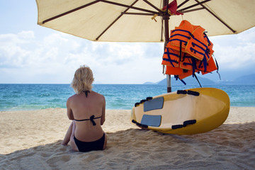 Woman in a swimsuit sitting on the sand on the beach. Surfboard and life vests on the...