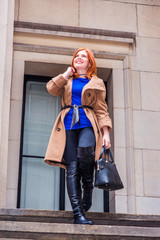 Obraz na płótnie Canvas American businesswoman traveling, working in New York, wearing long brown woolen overcoat, blue undershirt, black pants, long leather boots, holding hand bag, walking down stairs, looking up, smiling
