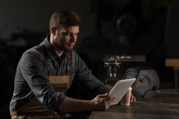 young male workman with paper cup of coffee using digital tablet