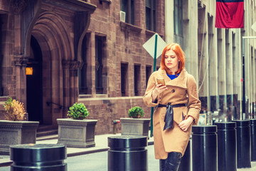 Young American businesswoman traveling in New York, wearing long brown woolen overcoat, sitting on metal pillar on narrow vintage street, looking down, texting on cell phone, taking work break..