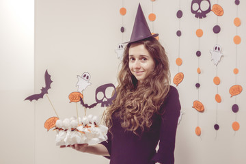 Portrait of charming young Witch in witches hat and black dress at Halloween Party.  Happy Halloween Witch holding candies like ghosts. Ghosts, circles, and skulls at background.