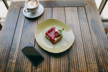 Close up of cake with raspberry sauce on plate, cup of cappuccino, mobile phone with blank empty screen lying on wooden table in coffee shop, cafe, restaurant. Overhead top view. Lifestyle concept.