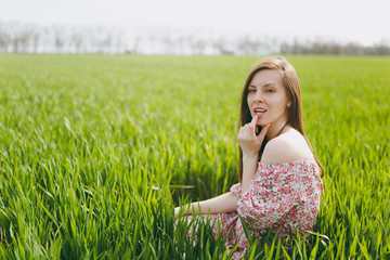 Young attractive pretty woman in light patterned dress sitting on grass keeping hand near mouth resting in sunny weather in field on bright green background. Spring nature. Lifestyle, leisure concept.