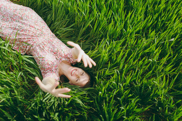 Young fun overjoyed beautiful woman in light patterned dress lying on grass raising hands up resting in sunny weather in field on bright green background. Spring nature. Lifestyle, leisure concept.