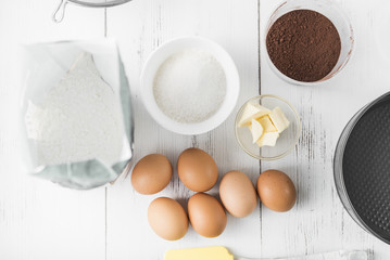 Ingredients for cooking: eggs, flour, sugar, cocoa powder, butter on a white wooden background, top view