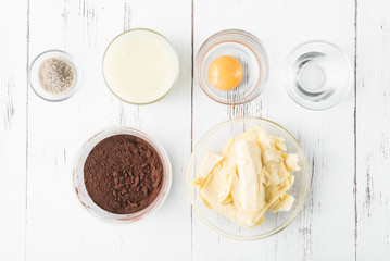 Ingredients for the preparation of cake cream: cocoa powder, butter, sugar, vanilla, yolk, condensed milk and water on a white wooden table, top view