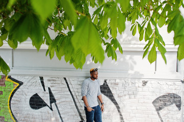 Stylish african american boy on gray sweater and black sunglasses posed at street. Fashionable black guy against graffity wall.