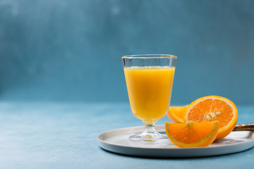 Glass of fresh orange juice with orange slices on white plate, blue background, cold summer refreshing nutritional beverage, blue orange color contrast, vitamin C healthy drink, copy space