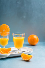 Glass of fresh orange juice with orange slices on white plate, blue background, cold summer refreshing nutritional beverage, blue orange color contrast, vitamin C healthy drink, copy space