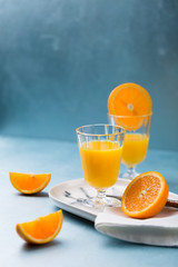 Glass of fresh orange juice with orange slices on white plate, blue background, cold summer refreshing nutritional beverage, blue orange color contrast, vitamin C healthy drink, copy space