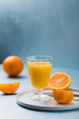 Glass of fresh orange juice with orange slices on white plate, blue background, cold summer refreshing nutritional beverage, blue orange color contrast, vitamin C healthy drink, copy space
