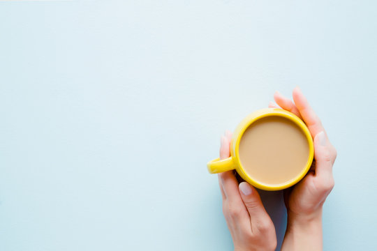 Woman's Hands Holding A Yellow Mug Of Coffee With Milk On Light Pastel Blue Table From Above. Wake Up With Morning Coffee. Empty Place For Inspirational, Motivational Text, Quote Or Logo. Top View.