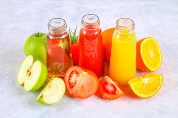 Bottles with fresh orange, apple, tomato juice on a gray concrete table. Fruits and vegetables around.