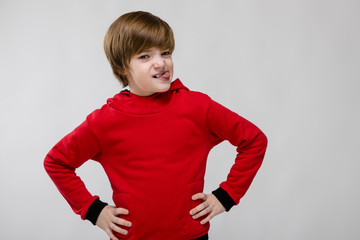 Cute confident caucasian little boy in red sweater holding hands on his waist on grey background