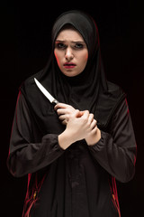 Portrait of beautiful desperate scared frightened young woman wearing black hijab holding knife in...