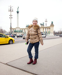 young little happy girl with hat on head is standing and posing happiness on square Hősök tere with yellow taxi background in Budapest and smiling