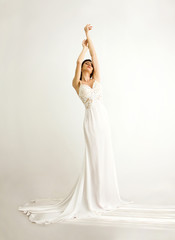 Naklejka premium young brunette sexy girl in a white wedding boudoir dress and jewellery in hairstyle is standing with hands on the top like swan princess on a white wall background 