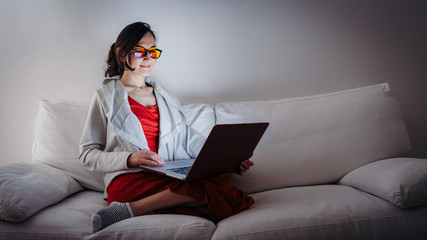 Young woman with blue light blocking glasses (yellow lenses) working with laptop on white sofa - Powered by Adobe