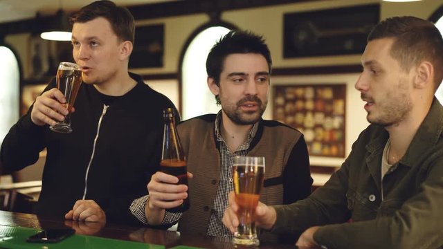 Male coworkers are having conversation in local bar. Young people are meeting outside workplace and communicating informally, clinking glasses and drinking beer.